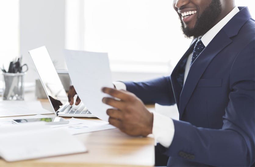 Professionnel souriant, assis à son bureau, travaillant sur son ordinateur portable et tenant un document à l'autre main