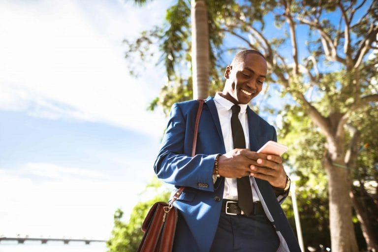 Professionnel, son sac de travail dans un bras, marchant à l'extérieur, souriant et regardant son téléphone portable