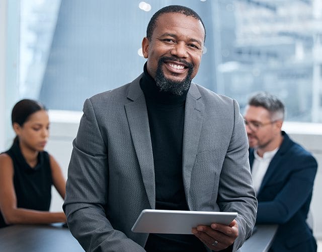 Professionnel confiant, fixant la caméra du regard, avec une tablette en main. Derrière lui, des collègues de travail