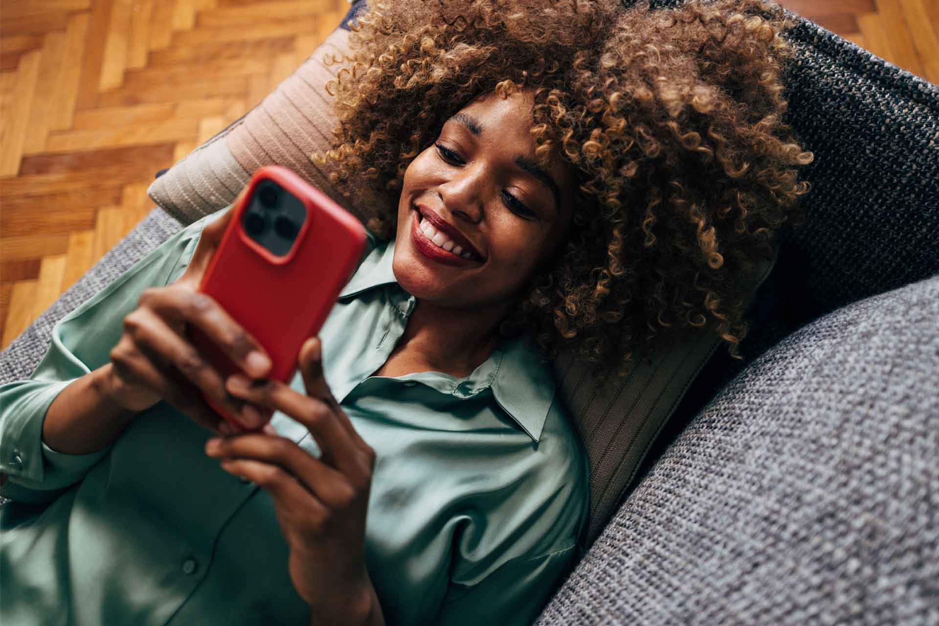 Jeune femme heureuse, détendue sur un divan, tenant son téléphone portable, utilisant avec joie les services bancaires en ligne de Capital Bank, Capital Bank Online