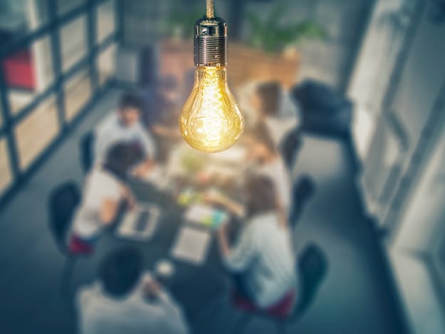 Salle de travail avec des collègues assis autour d'une table, une ampoule en premier plan symbolisant le brainstorming