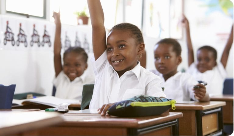 Jeunes enfants souriants et assis en classe avec les mains en l'air pour participer, symbolisant l'éducation