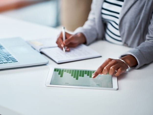 Illustration avec la main d'une femme consultant des graphiques financiers sur sa tablette tout en travaillant devant un ordinateur portable